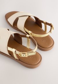 Sandales blanches et dorées avec une semelle plate marron. Elles comportent des brides croisées et une boucle pour un ajustement réglable. Texture en cuir lisse.