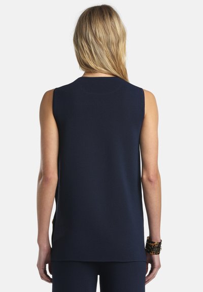 Personne blonde portant un haut sans manches bleu marine et un pantalon assorti, vue de dos sur un fond blanc uni.