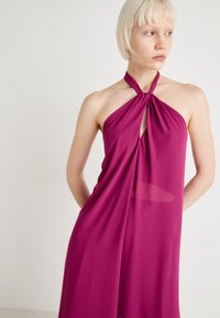 Magenta halter kleita, izgatavota no viegla auduma, ar atslēgas caurumu krūšu daļā un mezgla dizainu pie kakla.