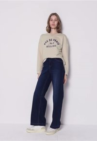 Beige sweatshirt met "RUE DE PARIS" opdruk, gecombineerd met donkerblauwe jeans met wijde pijpen. Model draagt lichtgekleurde sneakers en staat tegen een neutrale achtergrond.