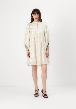 Lysfarget kjole med et sikksakkmønster, puffermer og rund utringning. Båret med svarte, strappy sandaler.