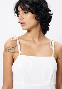 Weißes Leinenkleid mit zarten bestickten Blumenmustern, verstellbaren Bindebändern und einem quadratischen Ausschnitt. Glatte Textur.