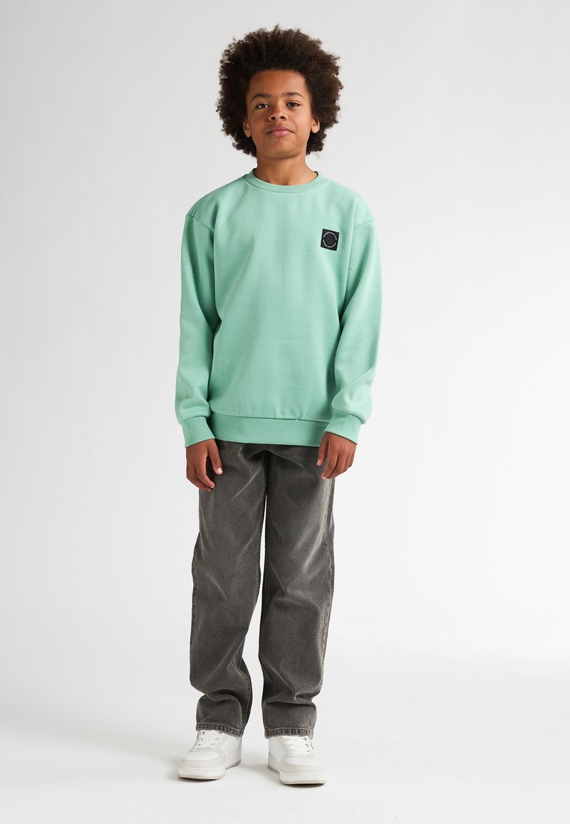 Sweatshirt vert menthe avec un patch logo noir, associé à un pantalon en denim gris et des baskets blanches. Coupe décontractée avec des poignets et un ourlet côtelés.