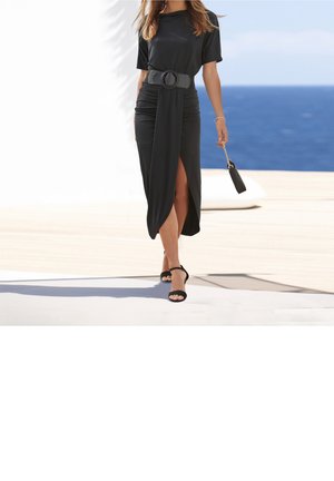 Robe maxi noire à manches courtes, avec des fronces, une fente latérale et une large ceinture texturée. Associée à des talons hauts noirs à brides et un petit sac.