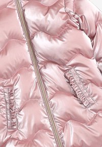 Lśniąca różowa pikowana kurtka z falistym przeszyciem, centralnym zamkiem błyskawicznym oraz dwiema pionowymi falbaniastymi kieszeniami po bokach.