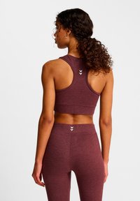 Mörkröd sportcrop top med racerback-design, texturerat tyg och tre vita chevron-accenter på baksidan. Figurnära modell.