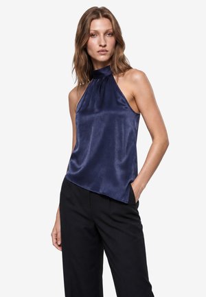 Vrouw draagt een mouwloze marineblauwe satijnen haltertop en zwarte broek, staat met één hand in haar zak tegen een effen achtergrond.
