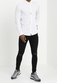 Hombre vistiendo una camisa blanca de botones con pequeños patrones negros, jeans ajustados rasgados negros y zapatillas gris plateadas, de pie contra un fondo blanco.