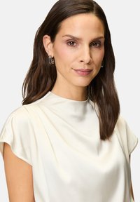 Cremefarbene Satinbluse mit hohem Ausschnitt und kurzen Ärmeln, die sanfte Drapierung und eine glatte Textur aufweist. Silbernene Creolen runden den Look ab.