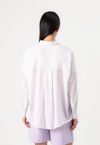 Camisa de botão listrada em roxo claro, com mangas longas, corte relaxado, pregas nas costas e uma barra suavemente curvada. Tecido de textura suave.