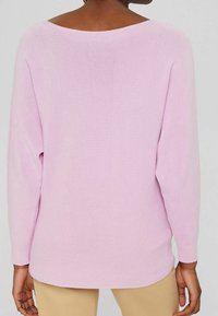 Rosa Pullover mit gerippter Textur, weitem Ausschnitt und überschnittenen Schultern. Längerer Saum hinten, getragen mit beigen Hosen. Einfaches und entspanntes Design.