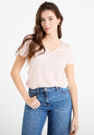 Junge Frau mit langen braunen Haaren, die ein hellrosa V-Ausschnitt-T-Shirt mit kurzen Ärmeln und hoch taillierte blaue Jeans trägt, steht mit einer Hand in der Tasche.