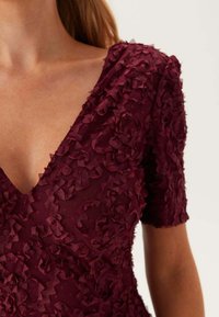 Burgundy klänning med V-ringning, prydd med texturerade blommönster och korta ärmar, tillverkad av lättviktsmaterial.