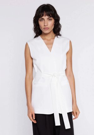 Kobieta o ciemnych, falowanych włosach, ubrana w białą, bezrękawnikową kamizelkę z paskiem i czarne spodnie, stojąca na tle jednolitego, jasnego tła.