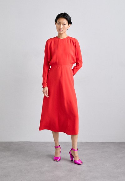 Vestido largo rojo de manga larga con un corte holgado, con un escote redondo y una cintura entallada. Combinado con zapatos de tacón alto brillantes en color rosa.