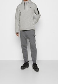 Grå huvtröja med svart logotyp, halvlång dragkedja och stor framficka; kombinerad med grå cargobyxor med sidofickor; svarta sportiga skor.