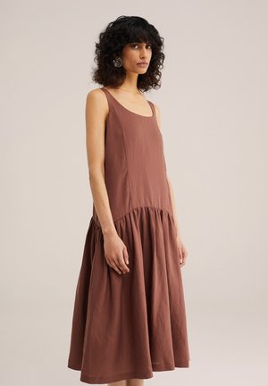 Naisel lokkis mustade juustega, kandes varrukateta pruuni kleiti kogutud seelikuga, seisab ühetaolise heleda beeži tausta ees.