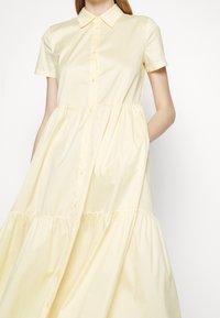 Femme portant une robe jaune pâle à manches courtes, boutonnée, avec un col et une jupe à volants, debout sur un fond blanc uni.