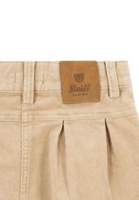Pantalones de pana beige con una etiqueta de cuero que presenta un logo de un oso y "Steiff desde 1880", mostrando pliegues y detalles de costura.