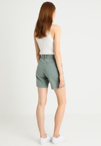Shorts en coton vert clair à coupe décontractée, cinq poches, et taille mi-haute. Associés à un débardeur blanc côtelé et des baskets blanches.