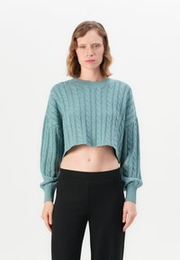 Kvinne iført en lyseblå cropped kabelstrikkegenser med lange ermer og svarte høytlivsbukser, stående mot en hvit bakgrunn.