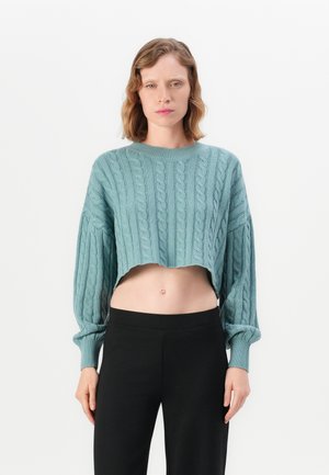 Kvinne iført en lyseblå cropped kabelstrikkegenser med lange ermer og svarte høytlivsbukser, stående mot en hvit bakgrunn.