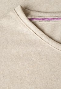Beige bomullstyg med en texturerad yta. Har en rundad krage och kontrasterande lila sömmar längs halsringningen.