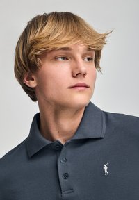 Polo gris oscuro con tejido texturizado, que presenta un logo bordado en blanco en el pecho izquierdo y tres botones en el cuello.