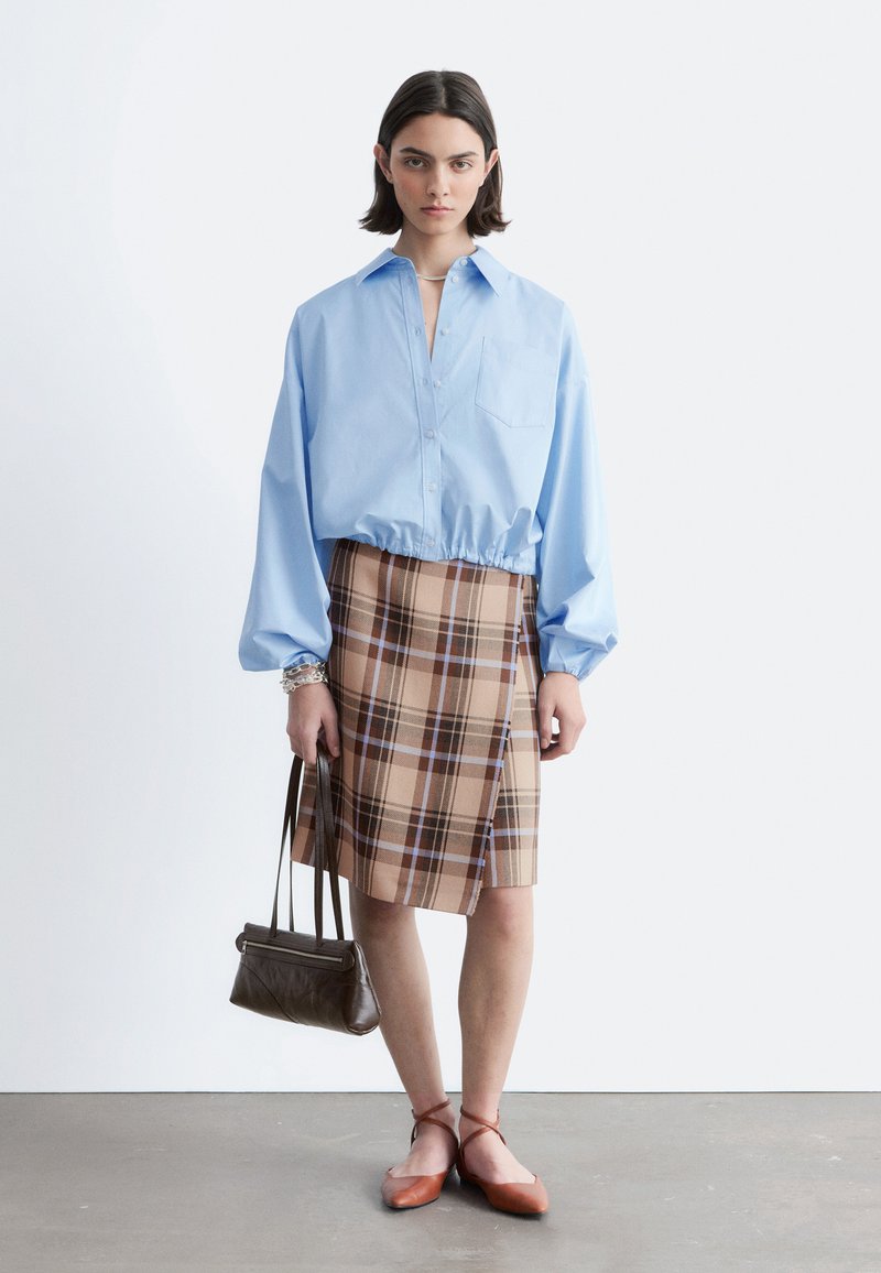 Jeune femme portant un chemisier bleu clair, une jupe à carreaux marron, des ballerines marron, tenant un sac à main marron, debout devant un fond blanc uni.