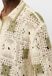 Camisa de crochet con botones en crema y verde oliva. Presenta patrones florales, hilo texturizado y diseño de tejido abierto con cuello.