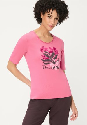 Kobieta o kręconych włosach, ubrana w różowy t-shirt z kwiatowym wzorem i ciemne spodnie, pozująca z jedną ręką na biodrze.
