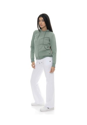 Une femme porte un sweat à capuche zippé vert sauge, un pantalon blanc et des baskets sur fond blanc.
