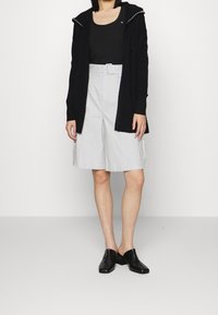 Cardigan nero a maniche lunghe, pantaloni corti grigi con tasche laterali e scarpe slip-on nere con texture. Design semplice e casual.