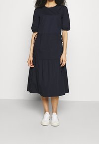 Mörkblå klänning med korta puffärmar, figursydd bodice, dragsnoddar i sidorna och en skiktad, knälång kjol. Matchad med vita sneakers.