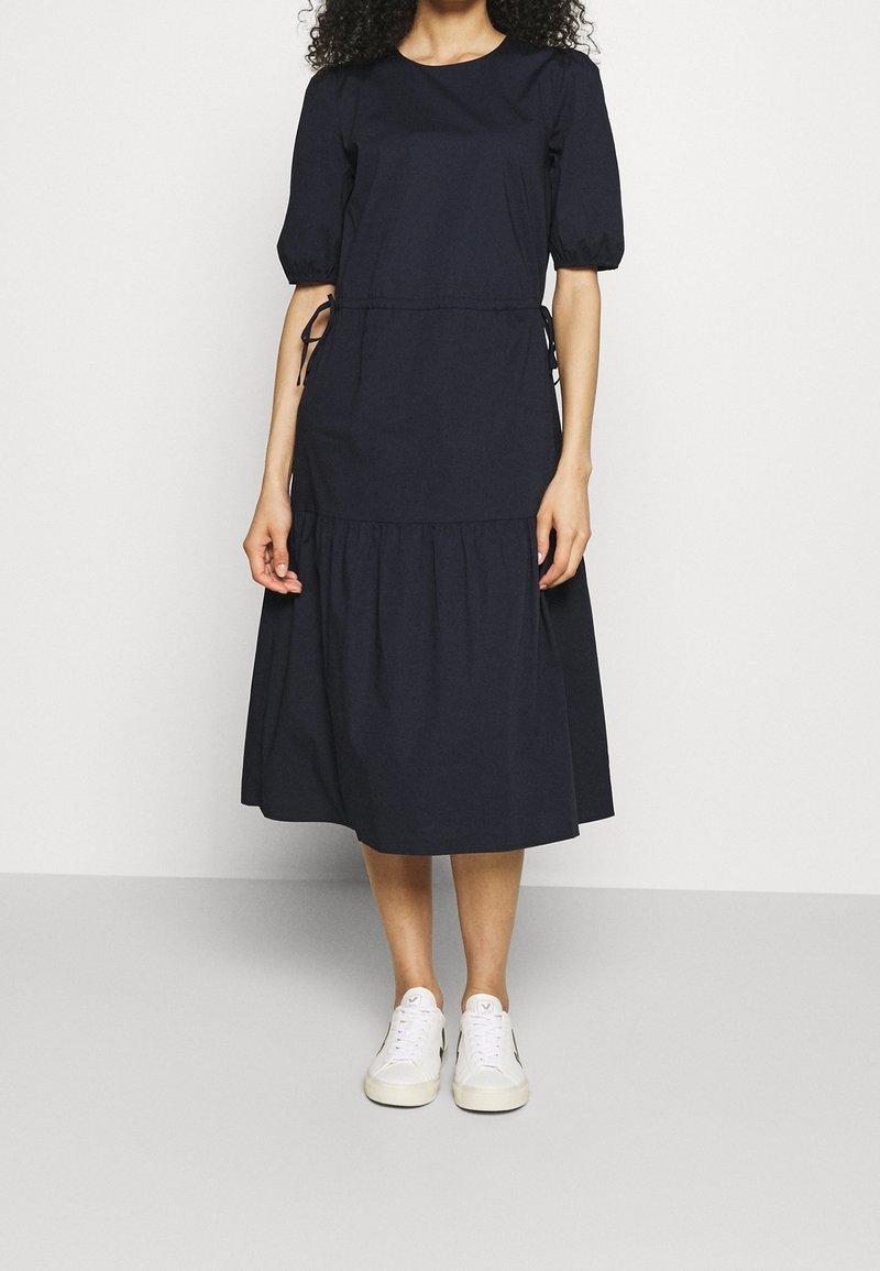 Mörkblå klänning med korta puffärmar, figursydd bodice, dragsnoddar i sidorna och en skiktad, knälång kjol. Matchad med vita sneakers.