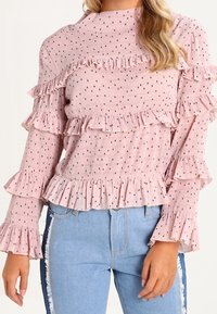 Světle růžová halenka s volánky a dlouhým rukávem s černými puntíky, s nařasenými detaily kolem výstřihu a rukávů, v kombinaci s džíny.