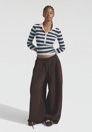 Jeune femme debout, les mains derrière le dos, portant un haut à manches longues rayé bleu et marron et un pantalon large ample marron foncé.