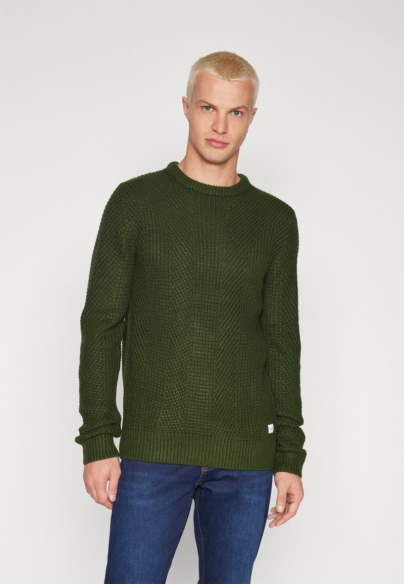 Dark green knitted sweater with a textured pattern, round neckline, and ribbed cuffs and hem. Worn with blue jeans.