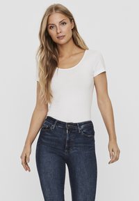 Bodysuit blanc à manches courtes avec encolure dégagée associé à un jean ajusté bleu foncé. Le jean présente une fermeture éclair et des détails de couture subtils.