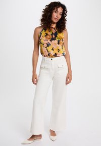 Květinový halter top s oranžovými, žlutými a černými vzory; design s vysokým krkem. Pár bílých širokých kalhot s předními kapsami a čistými liniemi.
