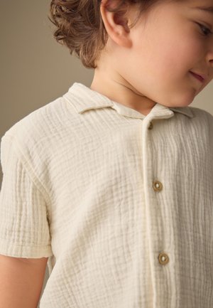 Jeune enfant aux cheveux courts et bouclés portant une chemise crème texturée à manches courtes boutonnée, de profil.