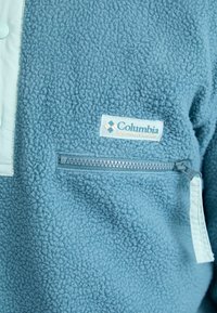 Lichtblauwe fleece trui met een ritszak, contrasterend lichtblauw paneel en Columbia logo patch. Zachte textuur en casual ontwerp.