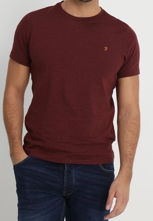 Mand iført en ensfarvet mørkerød T-shirt med korte ærmer og et lille orange logo på brystet samt mørkeblå jeans, stående med den ene hånd i lommen.