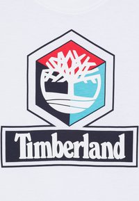 Camiseta blanca con un logo central que presenta un árbol estilizado, forma hexagonal, acentos en rojo y turquesa, y el texto "Timberland" en negrita debajo.