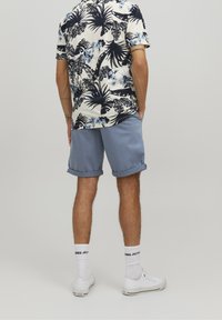 Camicetta floreale a maniche corte in crema e blu con foglie di palma, abbinata a pantaloni corti blu chiaro arrotolati e sneakers bianche con calzini bianchi.