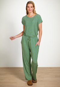 Vrouw staat binnen in groene en wit gestreepte losse broek en top met bruine slippers, glimlachend met één hand in de zak.
