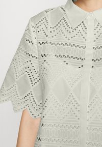 Blouse vert clair avec col pointu, présentant des motifs découpés complexes et des manches courtes festonnées, tissu texturé, et fermeture à boutons sur le devant.