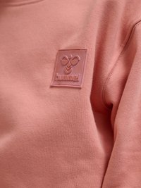 Close-up af en lyserød sweatshirt med et hævet gummipatch-logo med en stiliseret bi og ordet "hummel" på brystet.