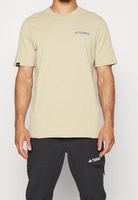 Beige bomulds-T-shirt med korte ærmer, der har en blå "Terrex"-logo på venstre bryst. Glat tekstur, afslappet pasform, rund hals.