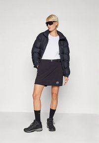 Melna pūkojoša jaka, balts T-krekls, melna svārki ar logotipu, melnas zeķes un melni pārgājienu apavi. Vienmērīga faktūra un cieši pieguļošs krojums.